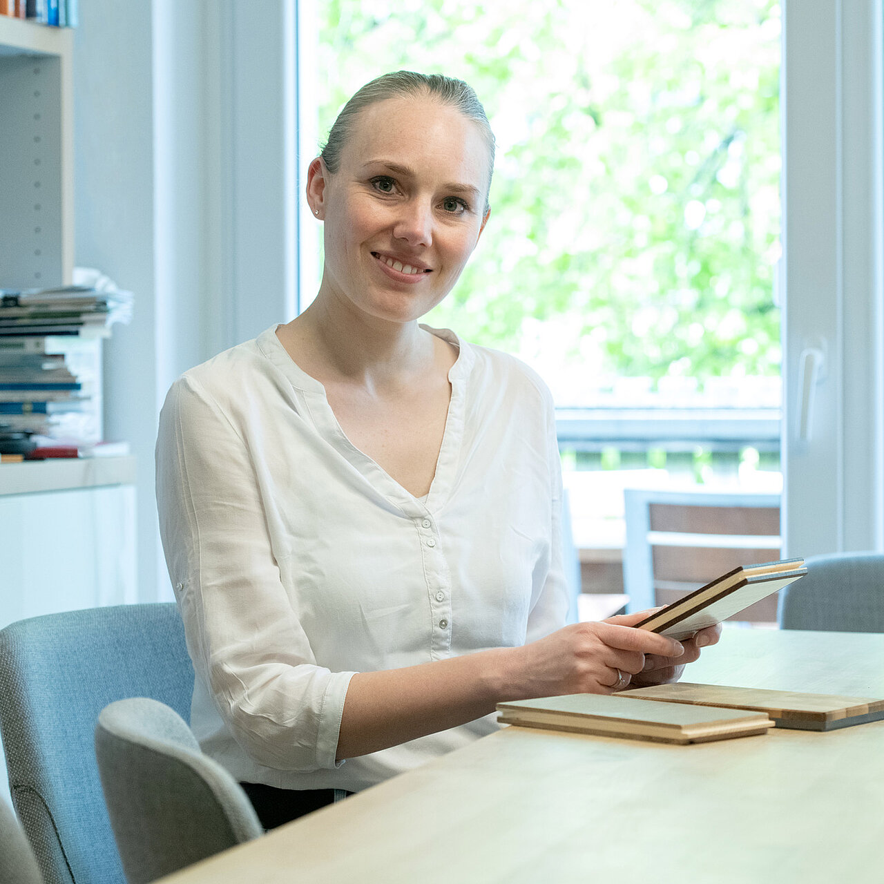 Lächelnde Person hält Holzmusterplatten am Tisch in hellem Büro